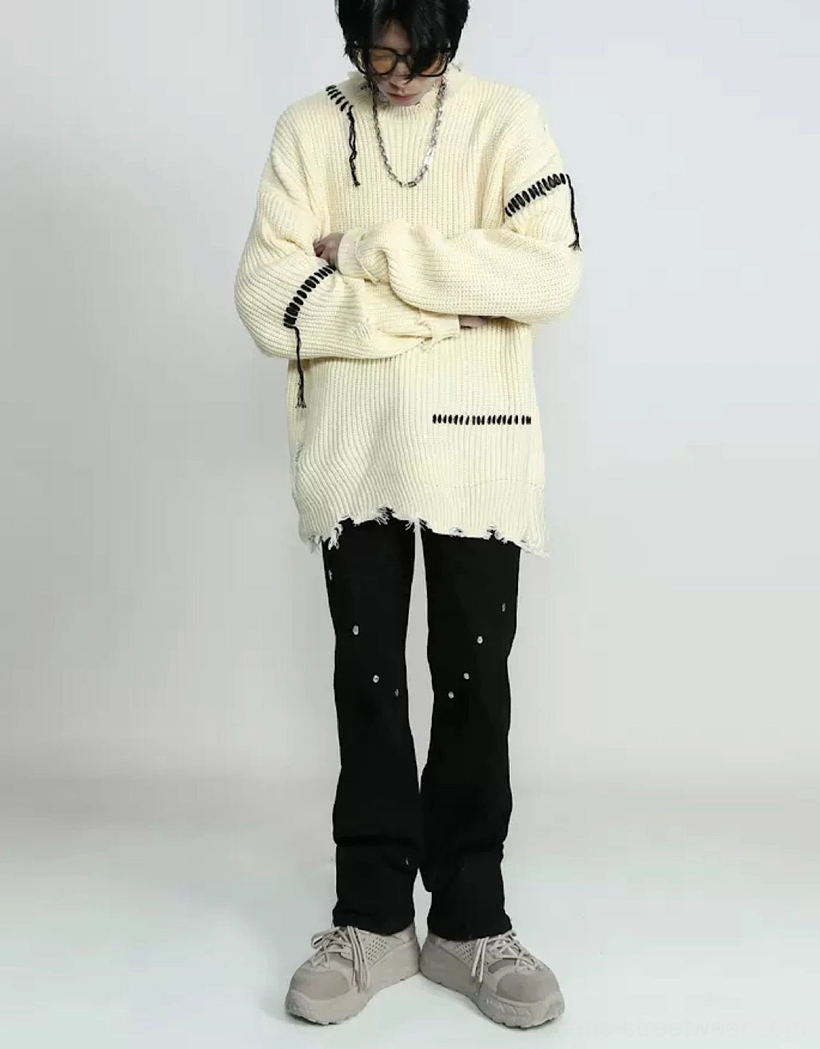Three-quarter front view of a cream apricot distressed knit sweater with decorative black stitching, long oversized sleeves, frayed hem, and silver necklaces