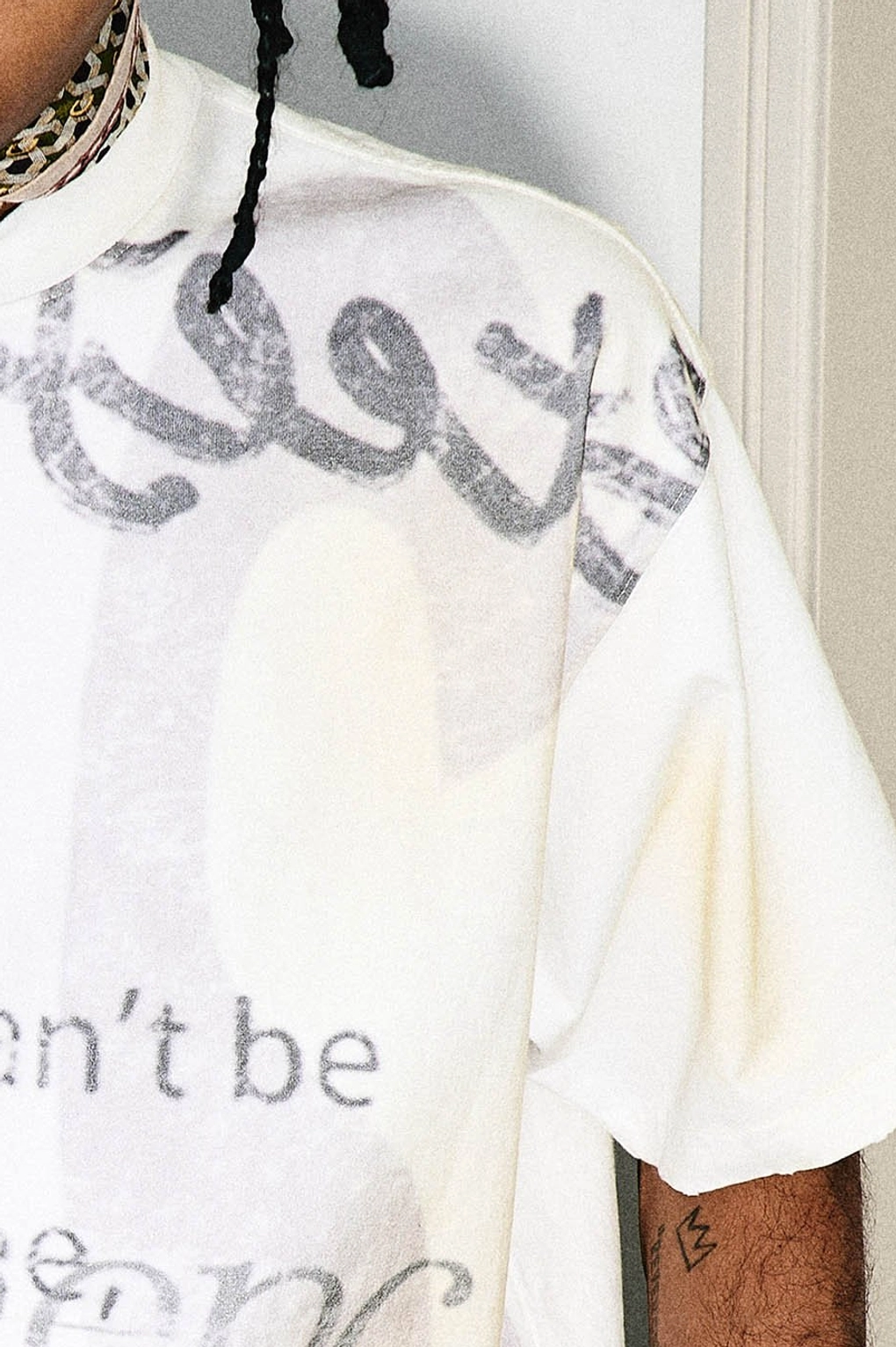 Back sleeve and shoulder detail of a cream streetwear T-shirt featuring large faded gray graffiti lettering