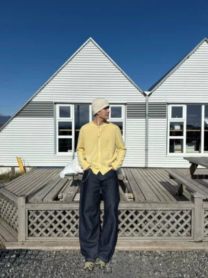 Creamy yellow henley knit cardigan full front view styled with a beige beanie and dark wide-leg jeans on a Scandinavian wooden deck