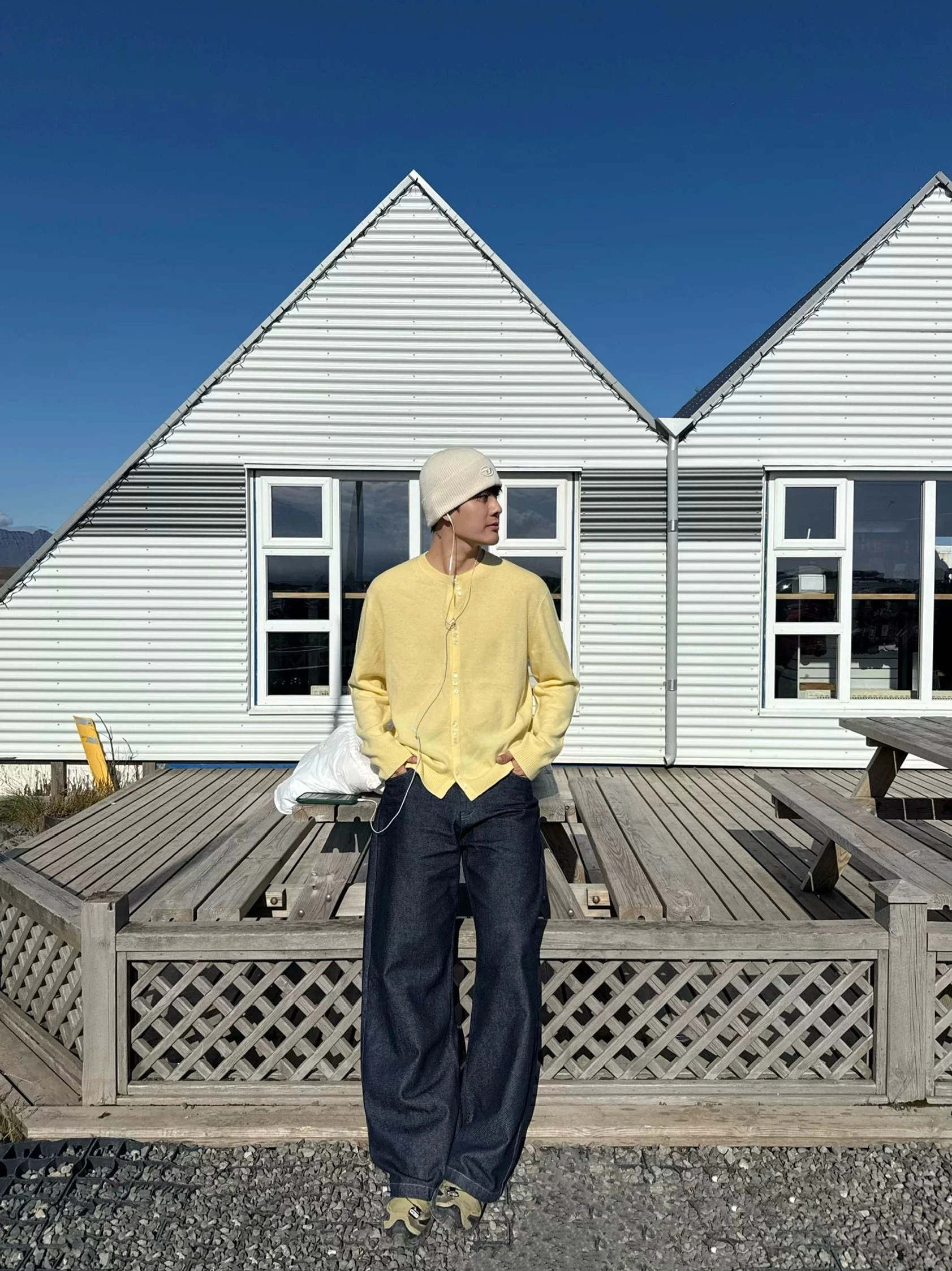 Creamy yellow henley knit cardigan full front view styled with a beige beanie and dark wide-leg jeans on a Scandinavian wooden deck