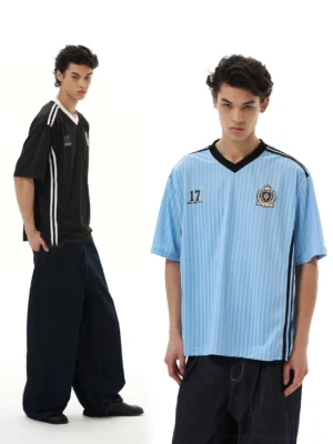 Light blue and black crested soccer jersey tees shown on male models with vertical pinstripes, V-neck collars, and wide-leg dark jeans