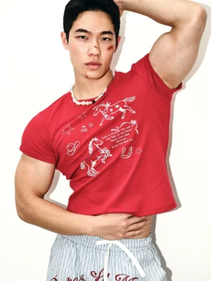 Front view of a muscular male model wearing a crimson red cropped Western rodeo graphic tee with white doodle artwork, striped lounge shorts, and a pearl beaded necklace