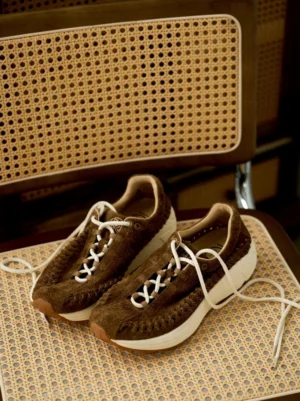 Dark brown handwoven suede runner sneakers displayed on woven rattan furniture with soft suede texture and thick off-white midsoles