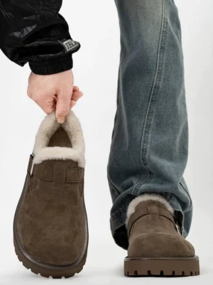 Dark brown shearling lined suede clog shown from the front with an adjustable metal buckle, thick non-slip lug sole, blue jeans, and black coat