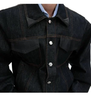 Close-up view of an oversized dark denim jacket showing leather-trimmed collar, crocodile-textured chest pocket panels, contrast stitching, and decorative metal floral buttons
