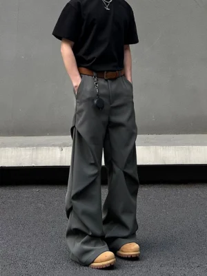 Dark graphite wide-leg suit trousers shown in a full front view with relaxed pooling legs, hidden side pockets, brown belt, and pendant detail in a concrete urban setting