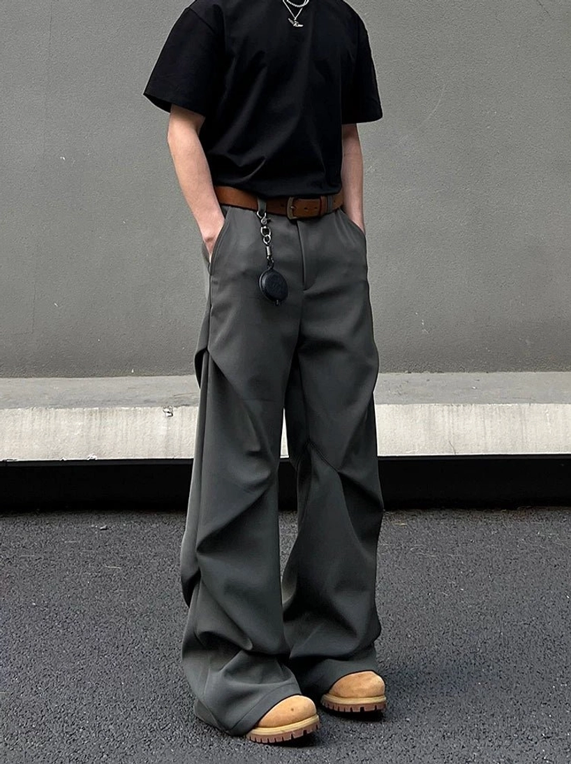 Dark graphite wide-leg suit trousers shown in a full front view with relaxed pooling legs, hidden side pockets, brown belt, and pendant detail in a concrete urban setting