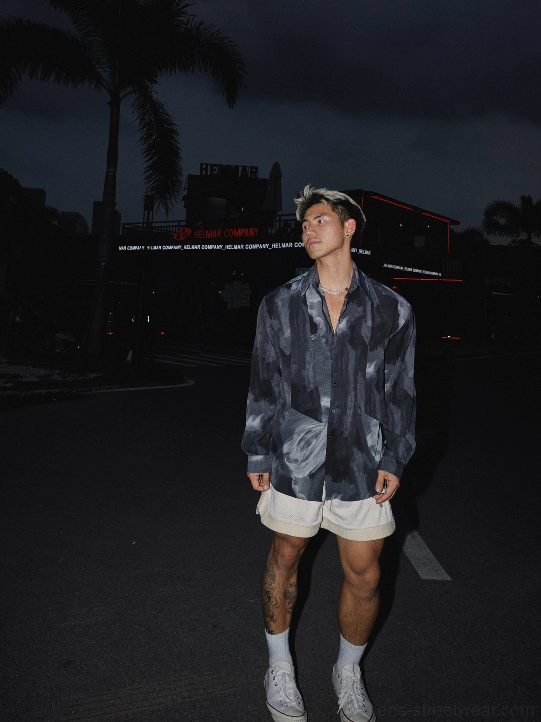 Full-body side view of a male model wearing a loose dark grey abstract satin shirt with casual shorts in a nighttime urban setting
