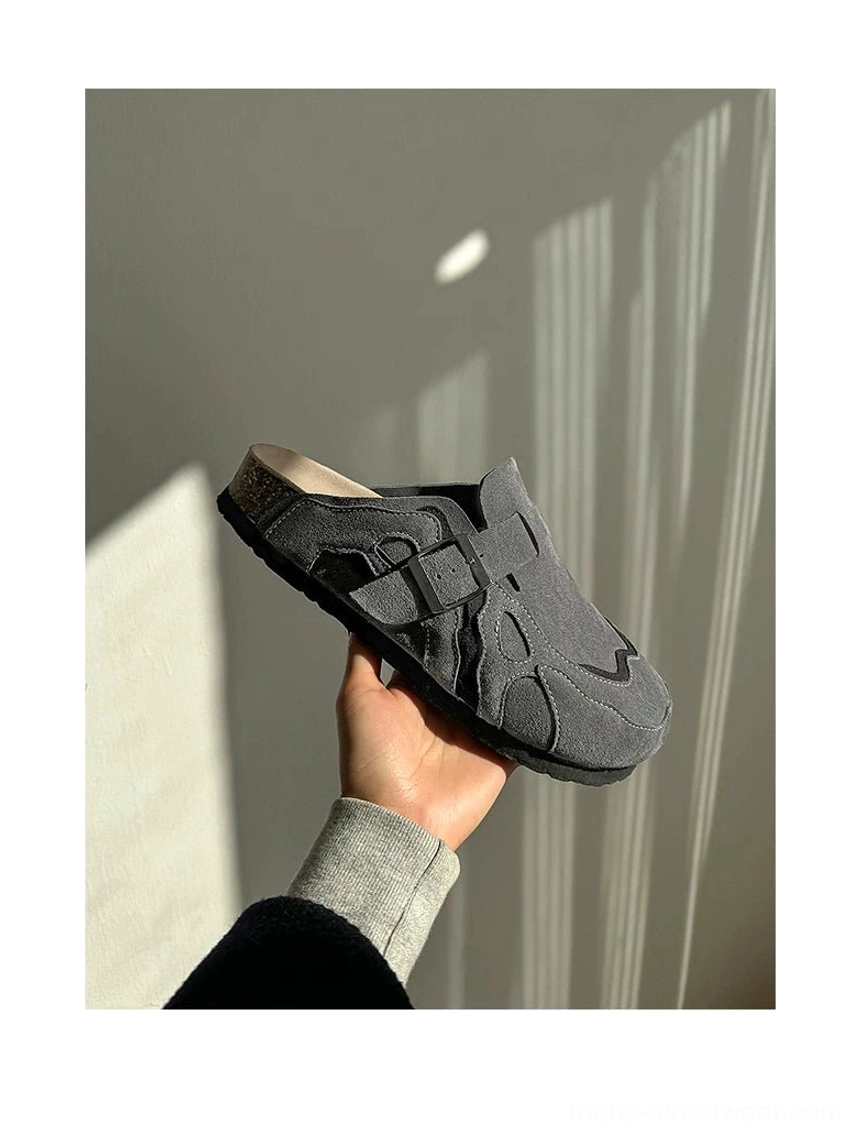 Handheld side close-up of dark grey suede clogs showing the curved overlays, buckle strap, and substantial composite sole
