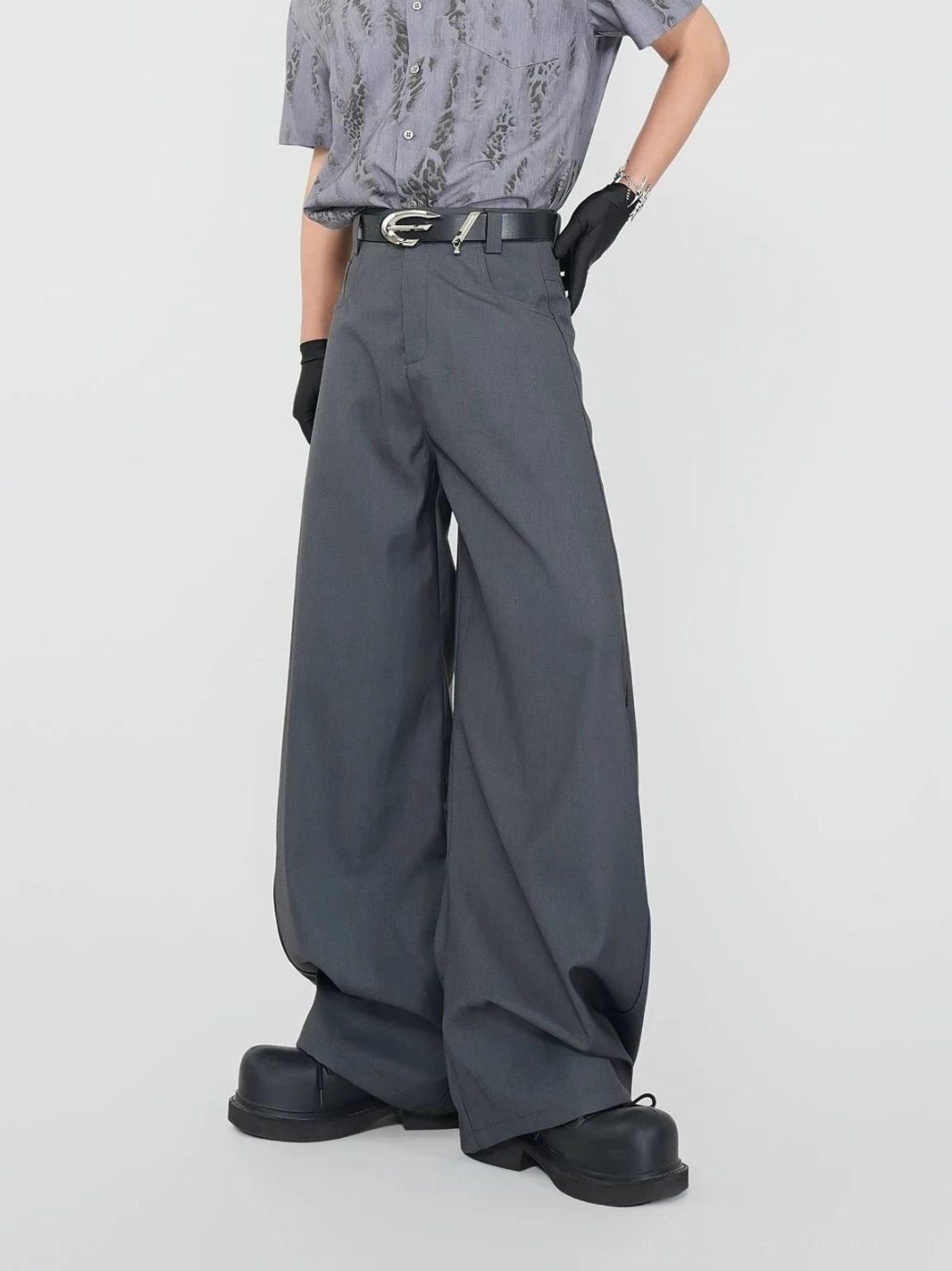 Three-quarter view of dark grey tailored wide-leg trousers showing the structured waistband, silver belt buckle, and fluid suiting fabric