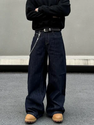 Dark indigo loose straight leg streetwear denim jeans shown from a low front angle against a minimalist concrete backdrop