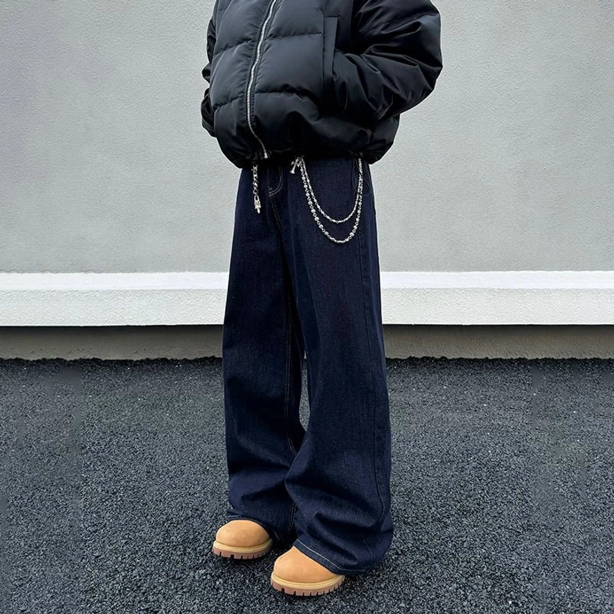 Low-angle front view of dark indigo wide-leg jeans with double metal waist chains, black cropped puffer jacket, and tan work boots