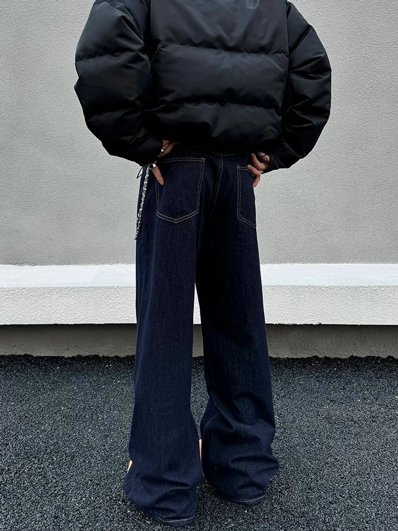 Back view of dark indigo wide-leg streetwear jeans styled with a black cropped puffer jacket, metal waist chain, and stacked hem on a rooftop