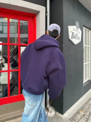Back view of a dark purple oversized zip-up hoodie showing its relaxed silhouette, thick fleece fabric and roomy hood on an urban street