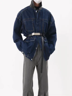 Dark wash oversized belted denim utility jacket shown from the front three-quarter view with high collar, cargo pockets, silver buckle waist belt, and relaxed streetwear fit