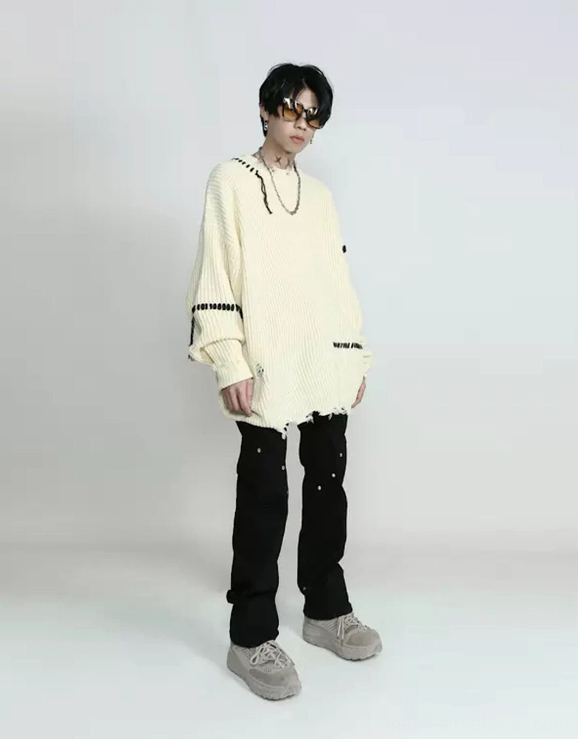 Full-body side view of a distressed apricot knit streetwear sweater styled with studded black trousers and grey suede sneakers
