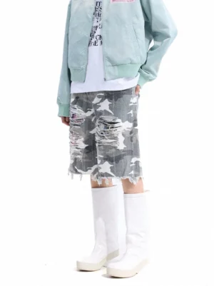 Front three-quarter view of distressed camouflage cutoff denim shorts with shredded thigh details, wide-leg streetwear fit, raw frayed hem, light blue bomber jacket, white tee, and platform boots