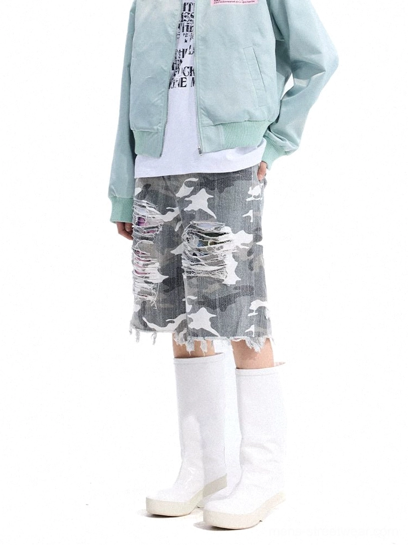 Front three-quarter view of distressed camouflage cutoff denim shorts with shredded thigh details, wide-leg streetwear fit, raw frayed hem, light blue bomber jacket, white tee, and platform boots