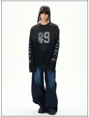 Front full-body view of a male model wearing a distressed black Force 09 graphic long sleeve tee with vertical sleeve lettering, wide-leg jeans, trapper hat, and work boots