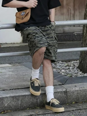 Model wearing black distressed leather canvas low-top sneakers with white socks and camouflage shorts in an urban streetwear setting