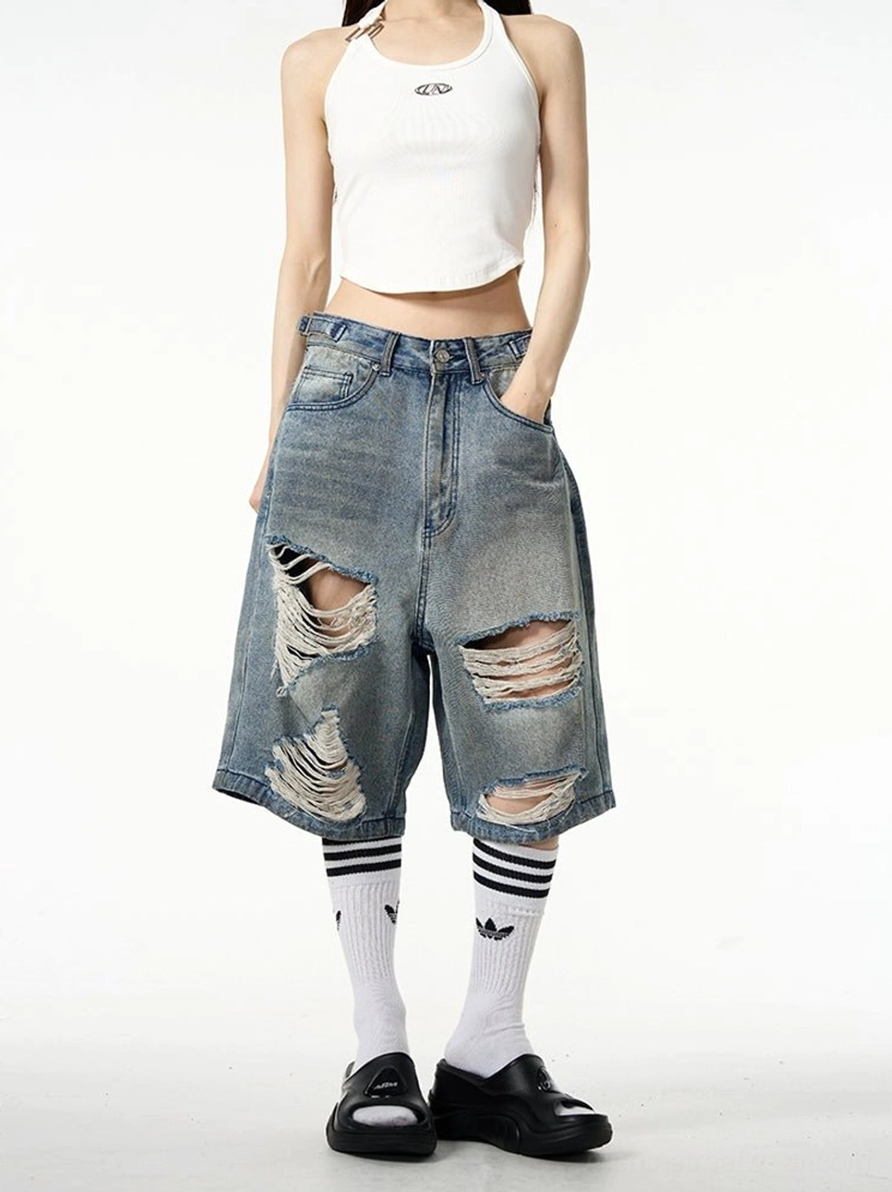 Front full-body view of oversized distressed ripped blue denim Bermuda shorts styled with a white cropped tank top, striped crew socks, and black loafers in a contemporary streetwear outfit