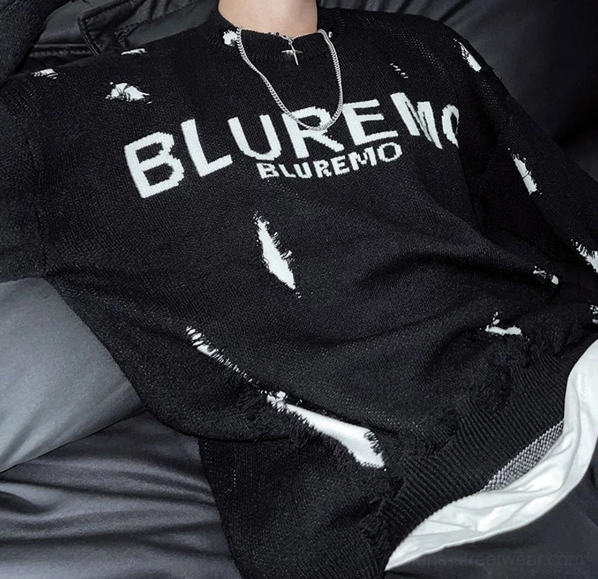 Close-up of black distressed shred-knit sweater chest lettering with visible knitted texture, frayed cutouts, and layered silver necklace styling