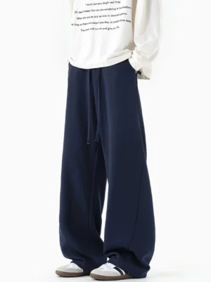 Navy blue draped wide-leg sweatpants shown from a front three-quarter angle with elastic drawstring waist, soft knit texture, oversized streetwear silhouette, white long-sleeve top, and casual sneakers