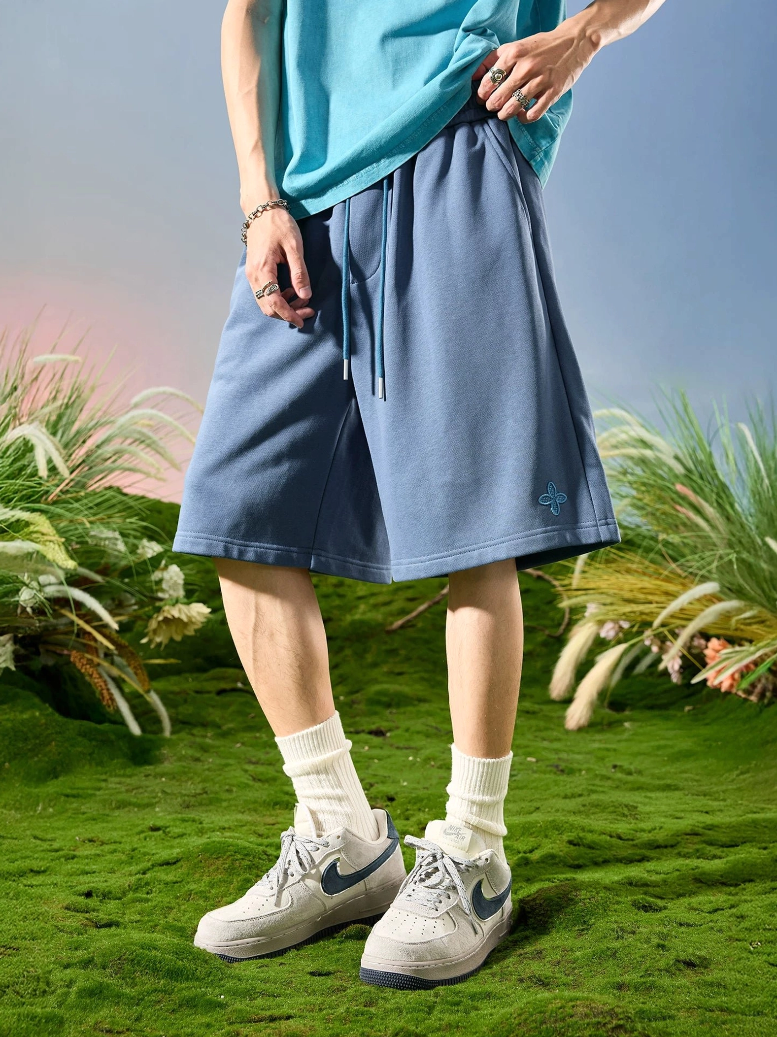 Front view of dusty blue clover embroidered sweat shorts with loose wide leg silhouette and comfortable cotton blend streetwear design