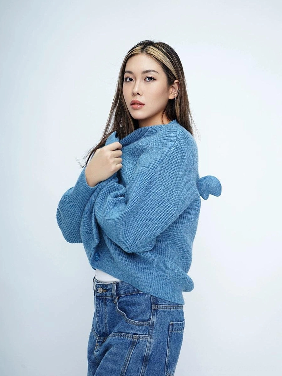 Female model wearing a dusty blue oversized knit cardigan with fuzzy texture, V-neck front, wing pom-pom back detail, and denim jeans