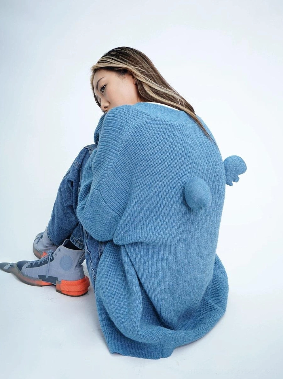 Dusty blue oversized fuzzy cardigan shown from a back three-quarter view with ribbed knit texture, wing pom-pom details, blue jeans, and sneakers