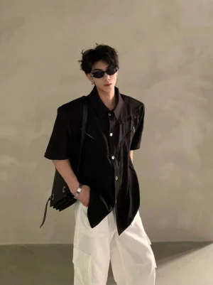 Black oversized short sleeve utility shirt with pointed collar, chest zipper detail, structured shoulders, sunglasses, white cargo pants, and black backpack