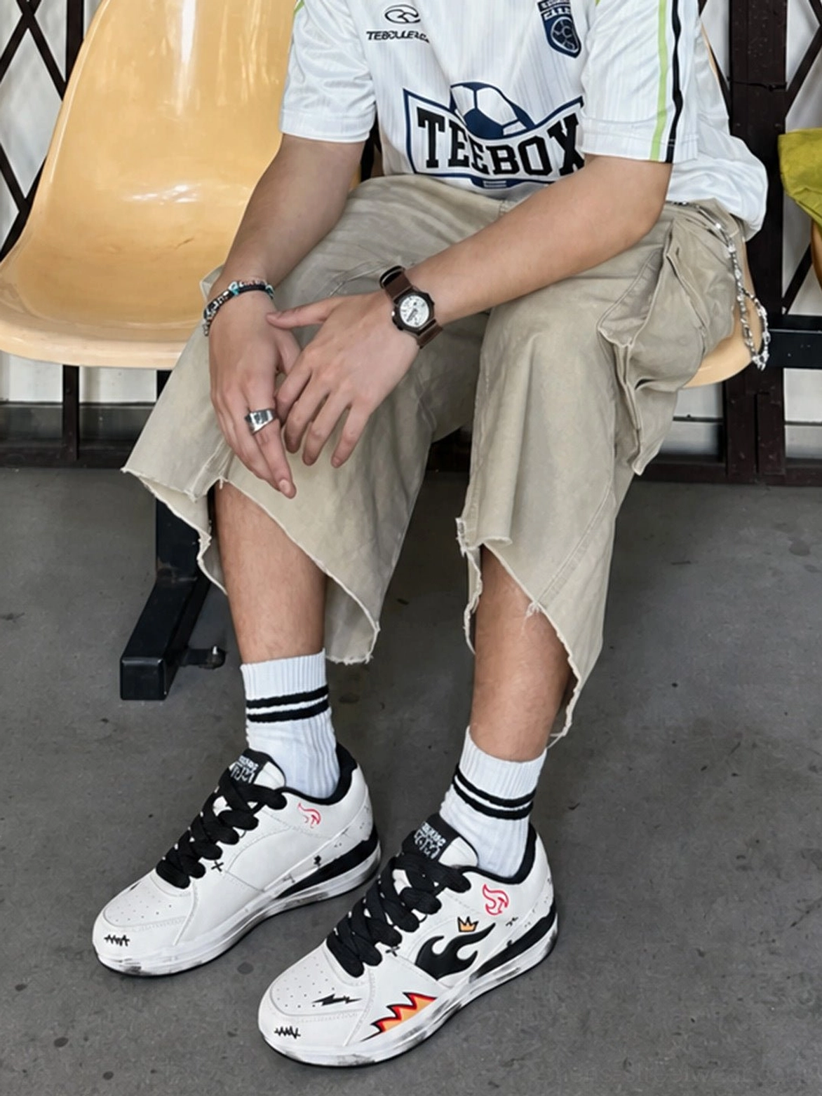 Model wearing flame graphic court sneakers with cropped khaki cargo pants, striped socks, and a football jersey in a casual streetwear setting