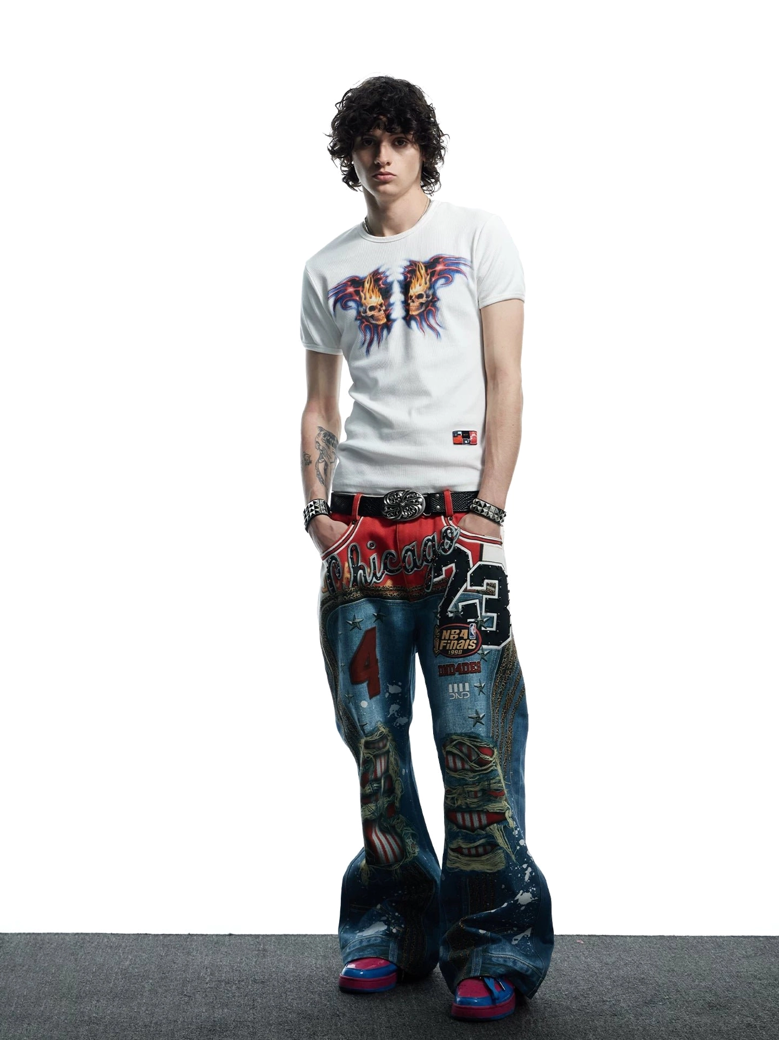 Three-quarter view of white flaming skull graphic baby tee styled with printed wide-leg denim and punk streetwear accessories