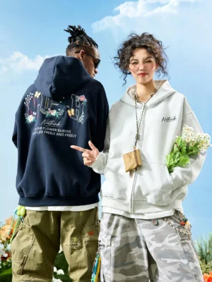 Floral embroidered oversized zip-up hoodie shown in dark navy and White Ash colorways against a bright blue sky with floral props
