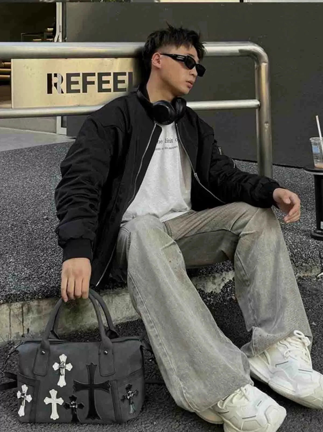 Black gothic crossbody satchel placed beside seated streetwear model in baggy jeans and sneakers