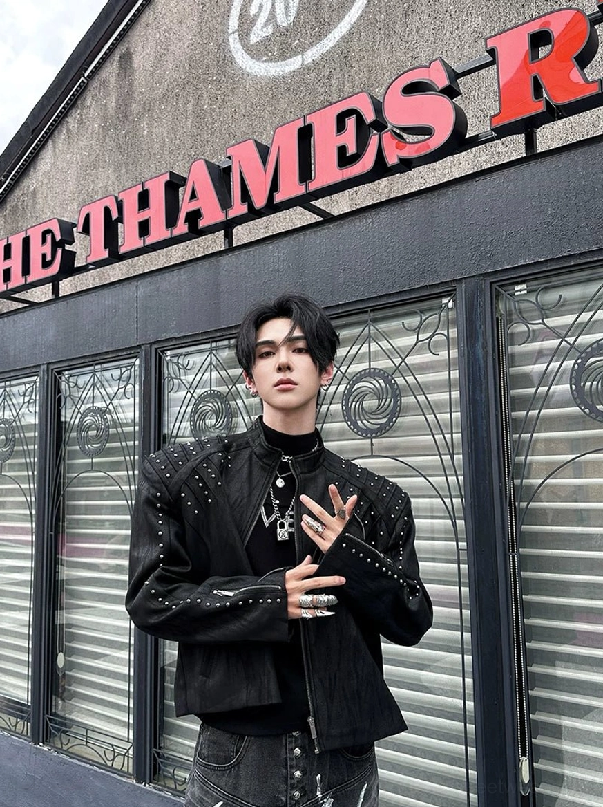 Male streetwear model in a studded gothic layered outfit with a fitted high neck top, biker jacket, graphic denim pants, and multiple silver necklaces