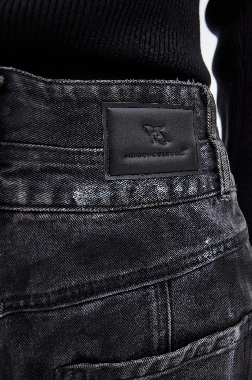 Close-up rear waist detail of graphite black distressed denim jeans showing leather patch belt loops washed texture and paint splatter finish