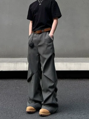 Graphite grey wide-leg draped trousers shown from a front three-quarter angle on a minimalist concrete street, styled with a brown leather belt and hanging pendant accessory