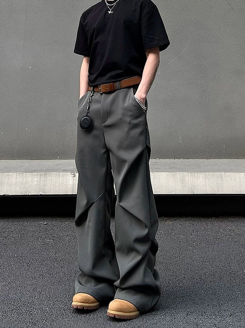 Graphite grey wide-leg draped trousers shown from a front three-quarter angle on a minimalist concrete street, styled with a brown leather belt and hanging pendant accessory