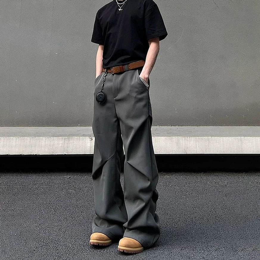 Graphite grey relaxed wide-leg trousers shown from a front three-quarter view with dramatic draped creases, black T-shirt, brown belt, pendant, and work boots
