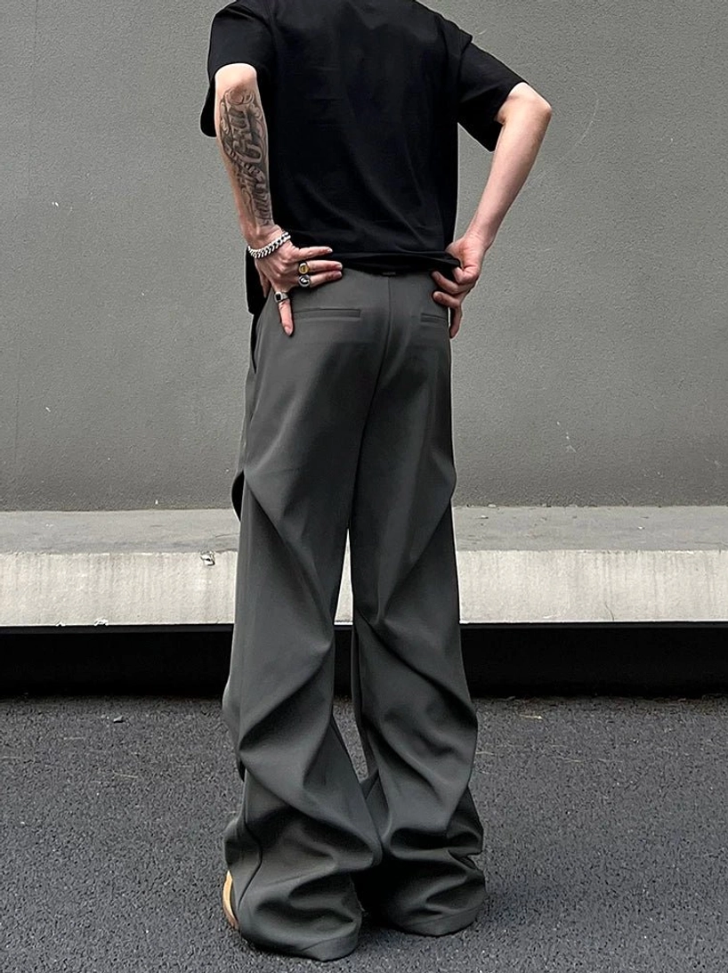 Graphite wide-leg draped trousers shown from the rear with relaxed folds, rear pockets, black T-shirt styling, and minimalist concrete streetwear backdrop