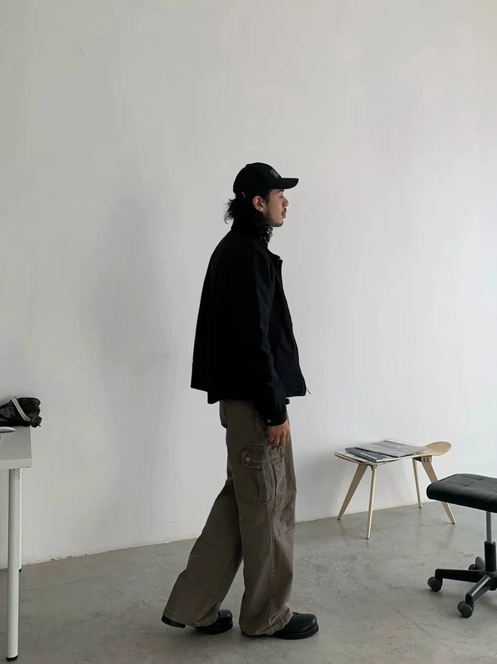 Side full-body view of gray khaki relaxed straight leg cargo pants with side cargo pockets styled with a black jacket and baseball cap