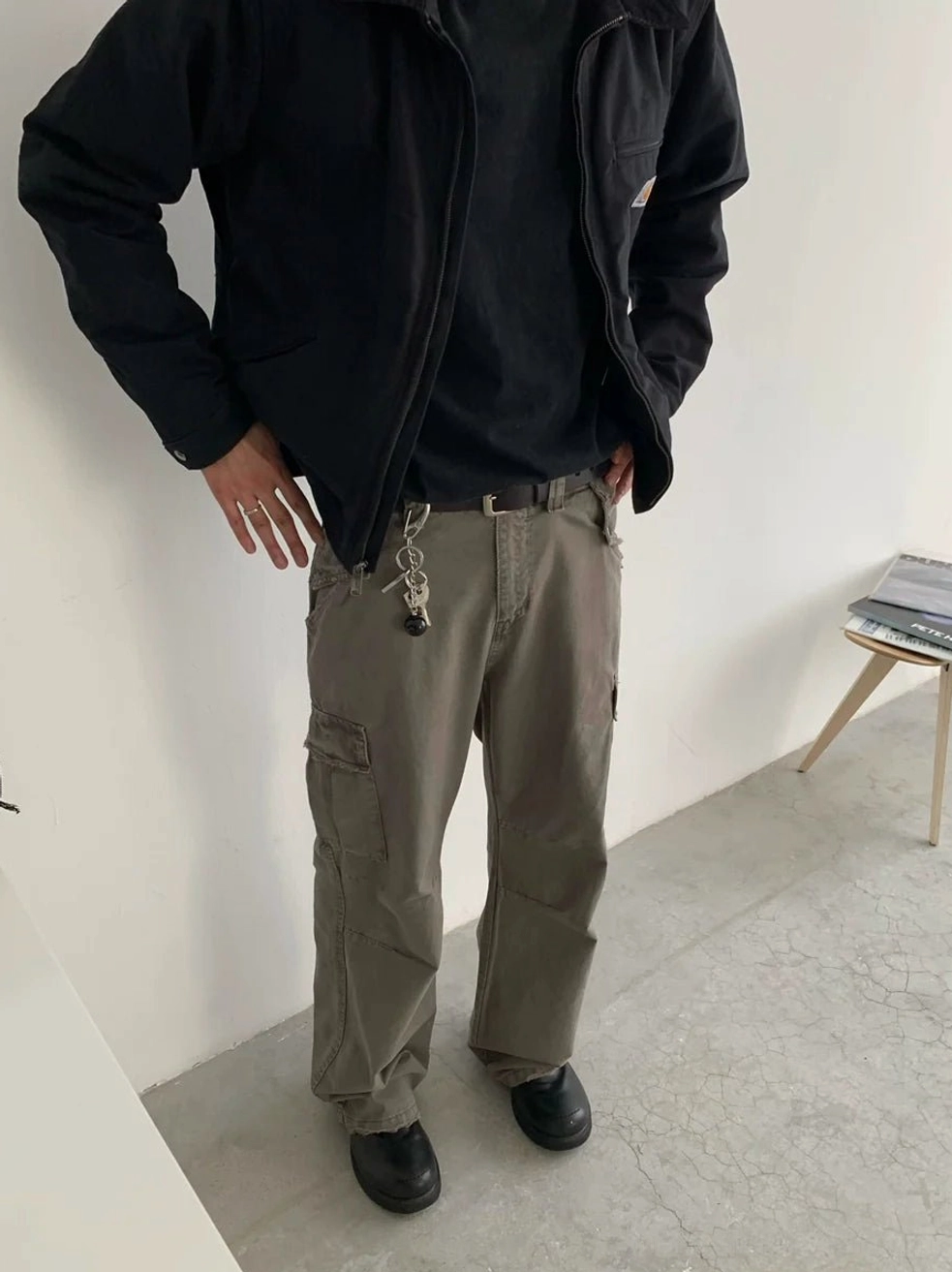 Front side view of gray khaki streetwear cargo pants with utility flap pockets and metal hanging ornament styled with a black cargo jacket