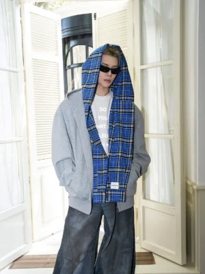 Male model wearing a gray oversized zip-up hoodie with blue plaid hood lining, white printed tee, sunglasses, and wide-leg washed denim in a modern streetwear outfit