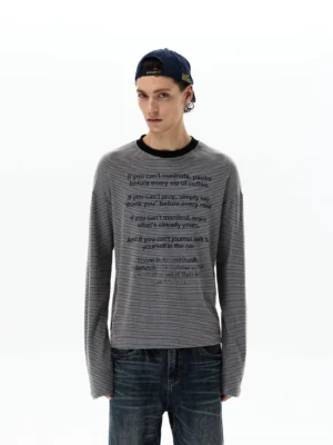 Front full body view of a gray striped mantra script long sleeve Korean streetwear tee styled with wide leg denim and a backward navy cap