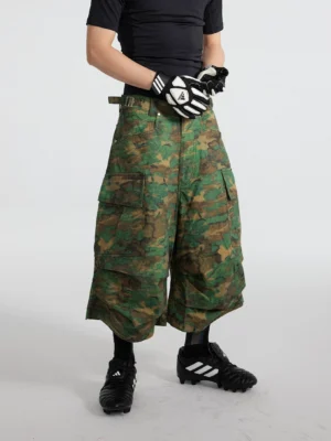 Model wearing green camouflage tiered cargo shorts with a wide-leg 3/4 silhouette, oversized pockets, sporty top, football boots, and streetwear styling