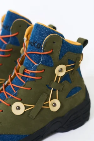 High-top green suede hiking boots with denim ankle collars, yellow inner lining, orange laces, and circular lace buckles shown in a side close-up