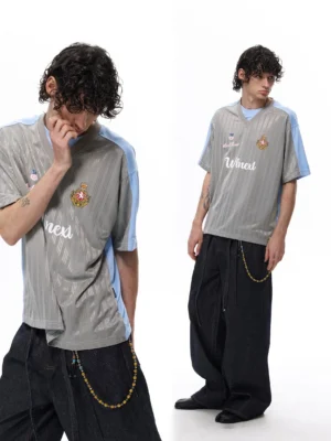 Close-up front view of a grey and pale blue retro soccer jersey T-shirt with subtle vertical stripe texture, crest detail, script lettering, V-neck collar, and modern streetwear styling