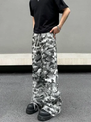 Front three-quarter view of grey abstract camouflage wide-leg cargo pants styled with a black T-shirt and chunky platform sneakers in a minimalist concrete streetwear setting