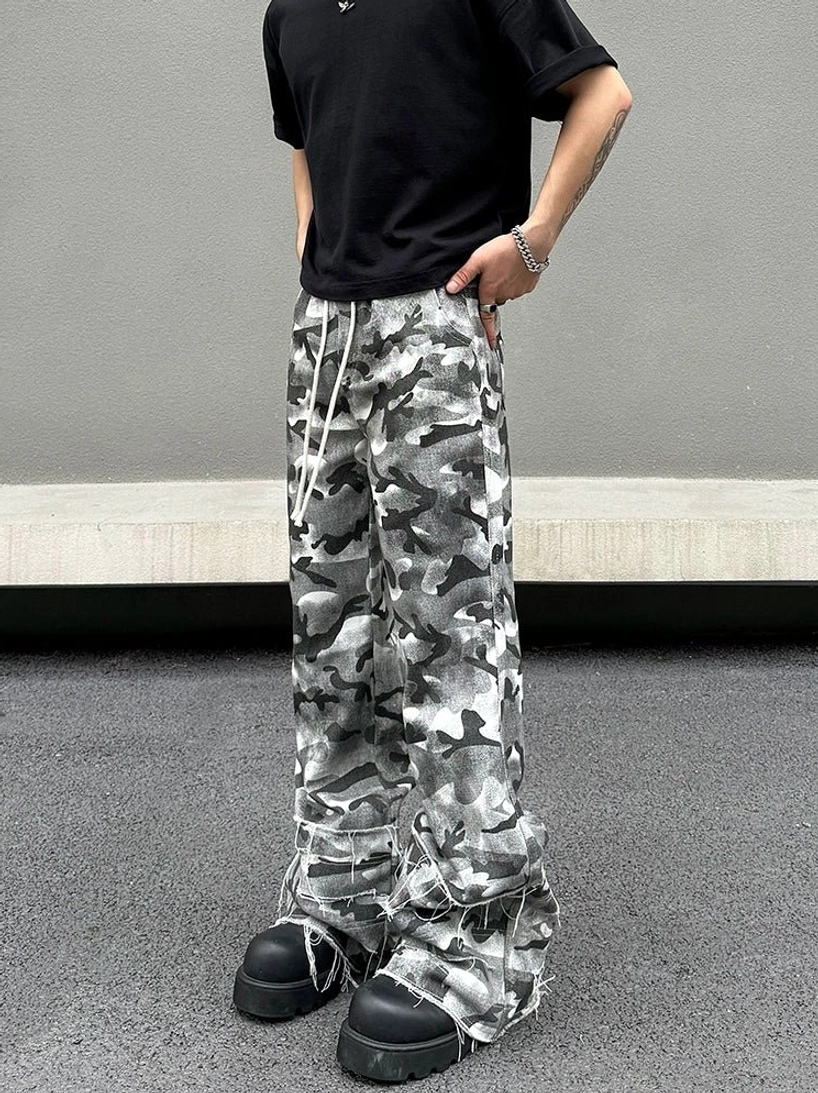 Front three-quarter view of grey abstract camouflage wide-leg cargo pants styled with a black T-shirt and chunky platform sneakers in a minimalist concrete streetwear setting